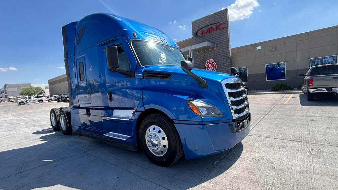 Blue Freightliner semi-truck cab in a dealership lot, front three-quarter view with chrome wheels and sleeper cab.
