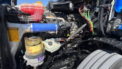 Truck engine bay with various hoses, wiring harnesses, and a blue silicone coolant hose. Visible components include a yellow oil/filters canister, coolant reservoir, power steering fluid reservoir, and a tangle of wiring harnesses. 