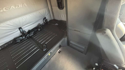 Interior of a Freightliner sleeper truck cab with a black ridged surface and seat area; visible text includes 'Scania' on light-colored upholstery. Image shows cabin floor area with seat and storage panel.