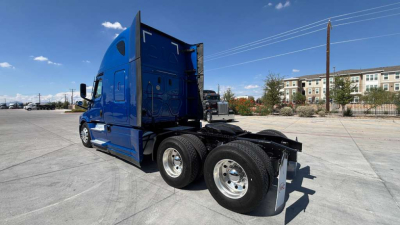 Truck cab and dual rear axles of a blue Freightliner semi-truck in a lot, side view, with chrome wheels and fifth wheel visible.