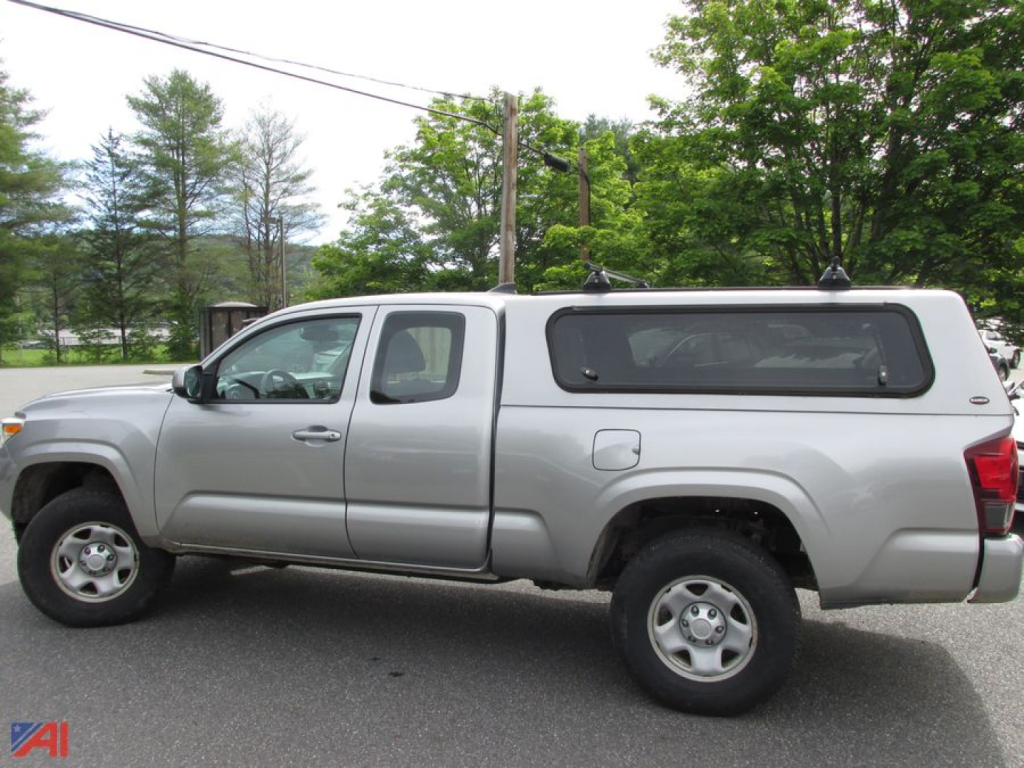 Side view of a 2018 Toyota Tacoma extended cab pickup truck with a gray color, black camper shell, and four-wheel drive, parked outdoors.