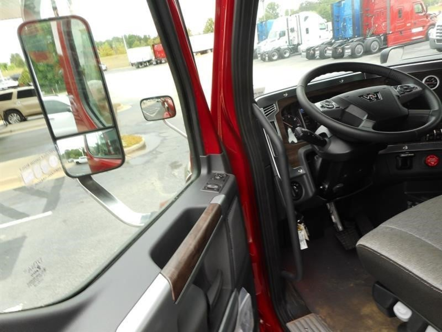 50% view of a Western Star 47X cab interior showing steering wheel and dashboard from the driver door, exterior red door frame, side mirror, and parking lot in background.