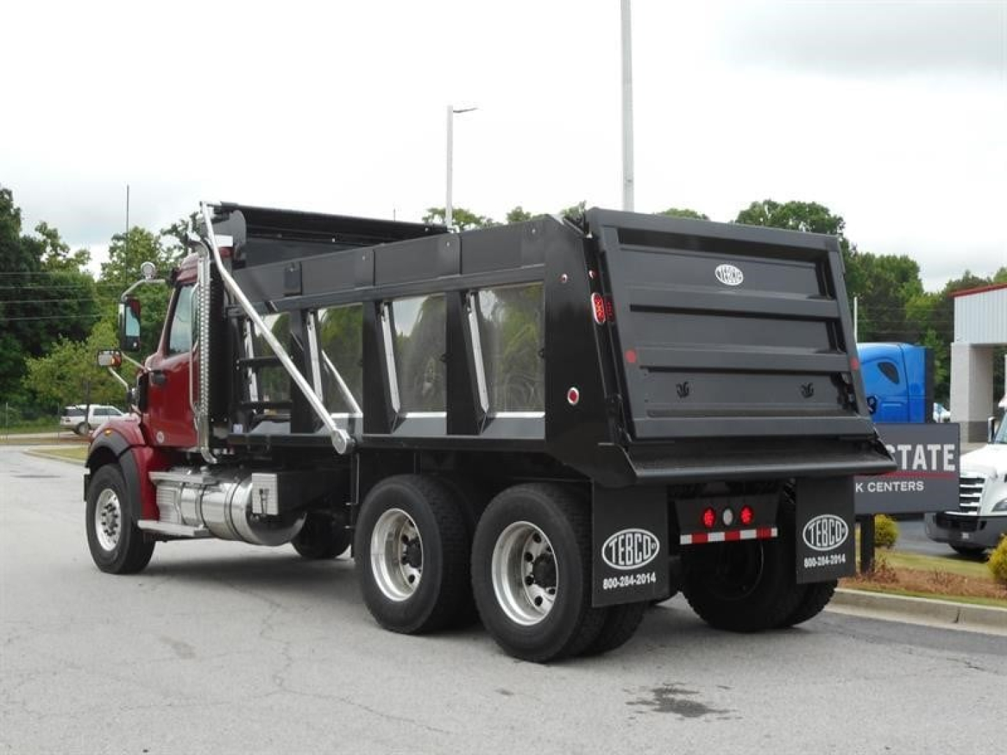 Western Star 47X dump truck rear view with black dump body and red cab, dual rear axles, chrome exhaust stacks, and White 'TBE' and other stickers on bumper.