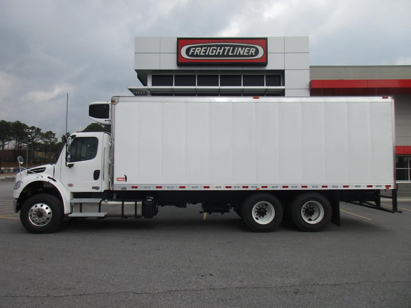 Freightliner M2 106 Plus refrigerated truck in front of dealership building with large Freightliner sign; cab is white, box is rectangular refrigerated cargo container.