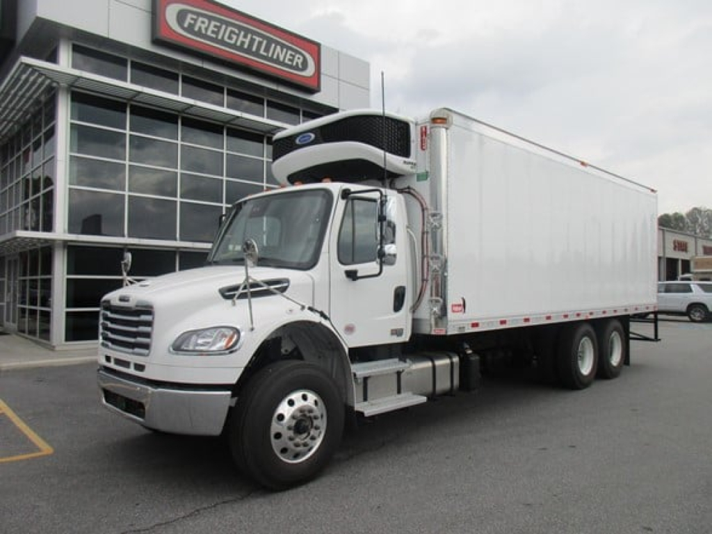 Freightliner M2 106 Plus refrigerated truck with a white cab and insulated box in front of a Freightliner dealership.