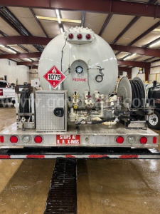 Tank truck rear showing 1075 placard, hazard panel, various valves, hoses, and storage access in a shop setting.