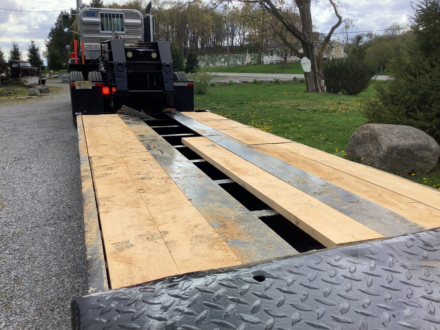 Two wooden planks on a flatbed trailer with a Mack RD8 truck in the background; the bed shows a split deck with openings and metal checker plate on the edge.