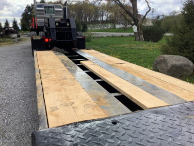 Two wooden planks on a flatbed trailer with a Mack RD8 truck in the background; the bed shows a split deck with openings and metal checker plate on the edge.