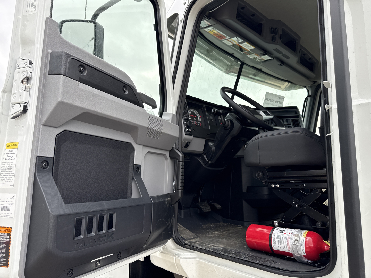 Interior of a new 2026 Mack PI64T daycab showing open driver door, steering wheel, dashboard, and a red fire extinguisher on the floor.