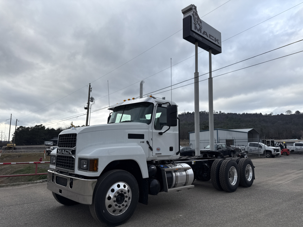 Truck chassis showing Mack PI64T cab over engine with dual rear axles and front grille, white exterior, long wheelbase, in dealership lot.
