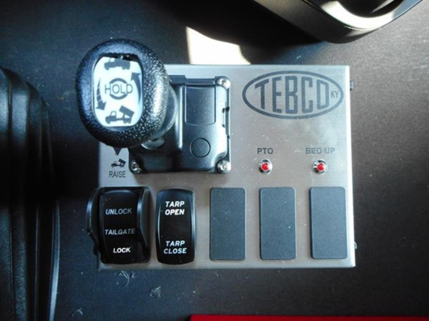 TEBCO KY badge panel with PTO indicator lights and control switches in a truck cab; includes “TAILGATE LOCK” and “TARP OPEN/CLOSE” labeled switches; circular hold/shift knob visible.