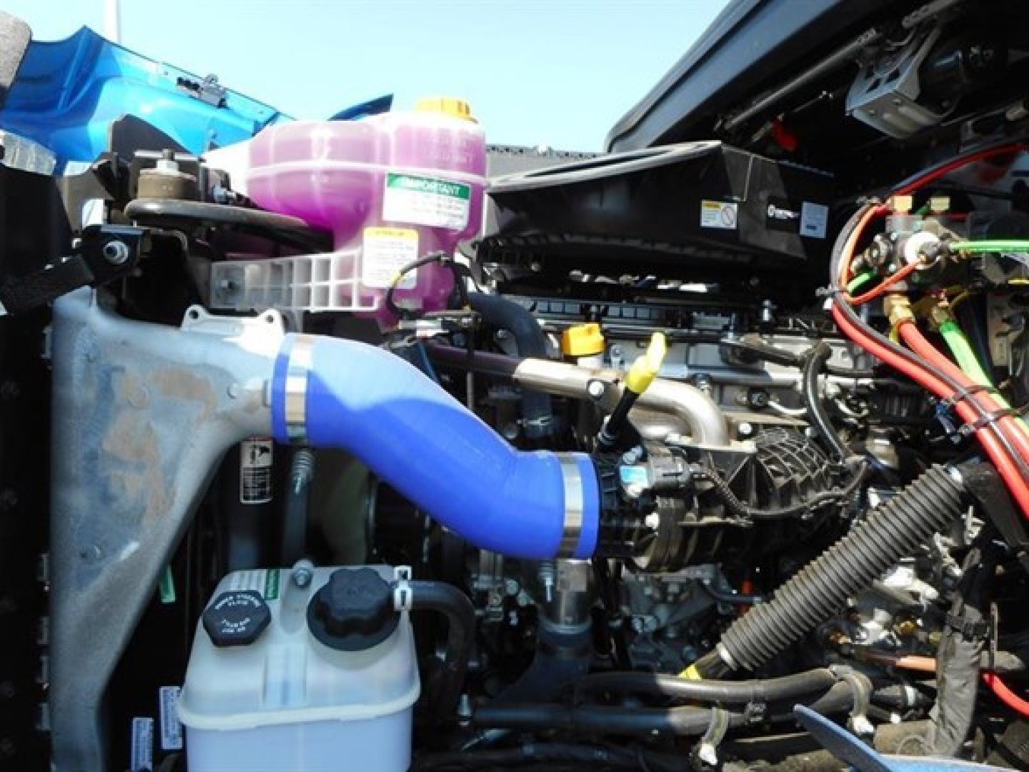 Vehicle engine bay of a 2026 Western Star 47x dump truck showing blue intercooler pipe, coolant reservoir, purple coolant/washer bottle, and various hoses and wiring