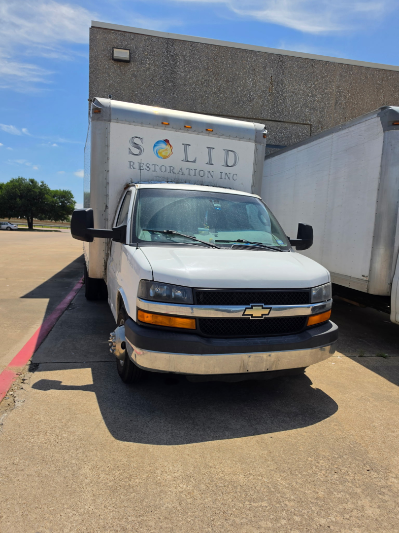 Chevrolet Express 3500 box truck front view showing cab and cargo box with 'SOLID RESTORATION INC' sign on nearby trailer; headlights and grille visible.