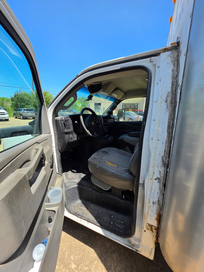 Chevrolet Express 3500 cargo van interior view from open driver door showing steering wheel, dashboard, and seat; exterior door frame with some rust and weathered area.
