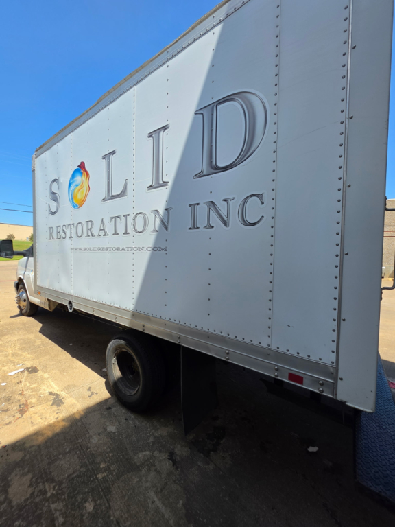 Box truck with SOLID RESTORATION INC logo on side, showing text SOLID RESTORATION INC and website, with rear wheel and box side.