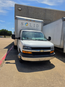 Chevrolet Express 3500 box truck front view showing cab and cargo box with 'SOLID RESTORATION INC' sign on nearby trailer; headlights and grille visible.