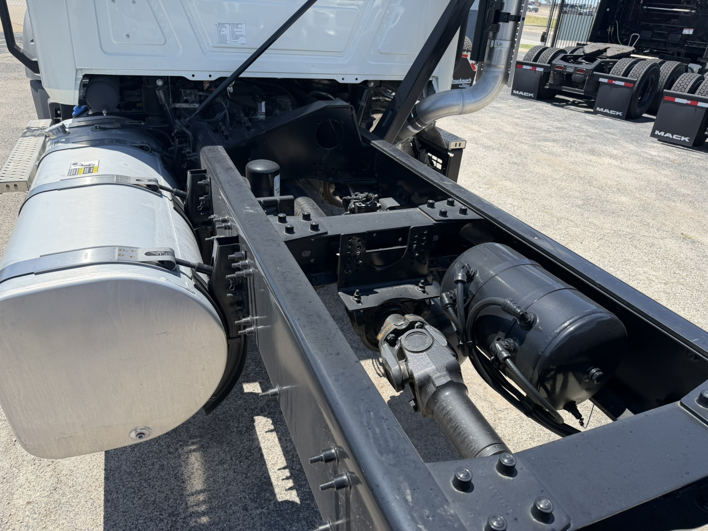 Chassis view of a 2025 Mack GR64F cab and chassis showing the fuel tank, frame rails, drive shaft, and engine bay with various hoses and components.