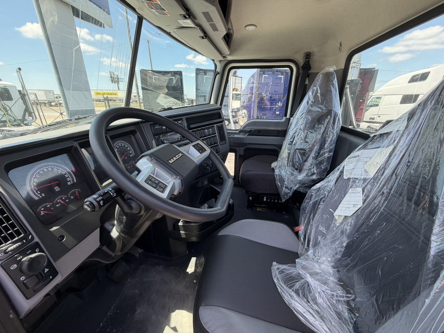Cab and chassis interior of 2025 Mack GR64F showing steering wheel, dashboard gauges, and plastic-wrapped driver and passenger seats
