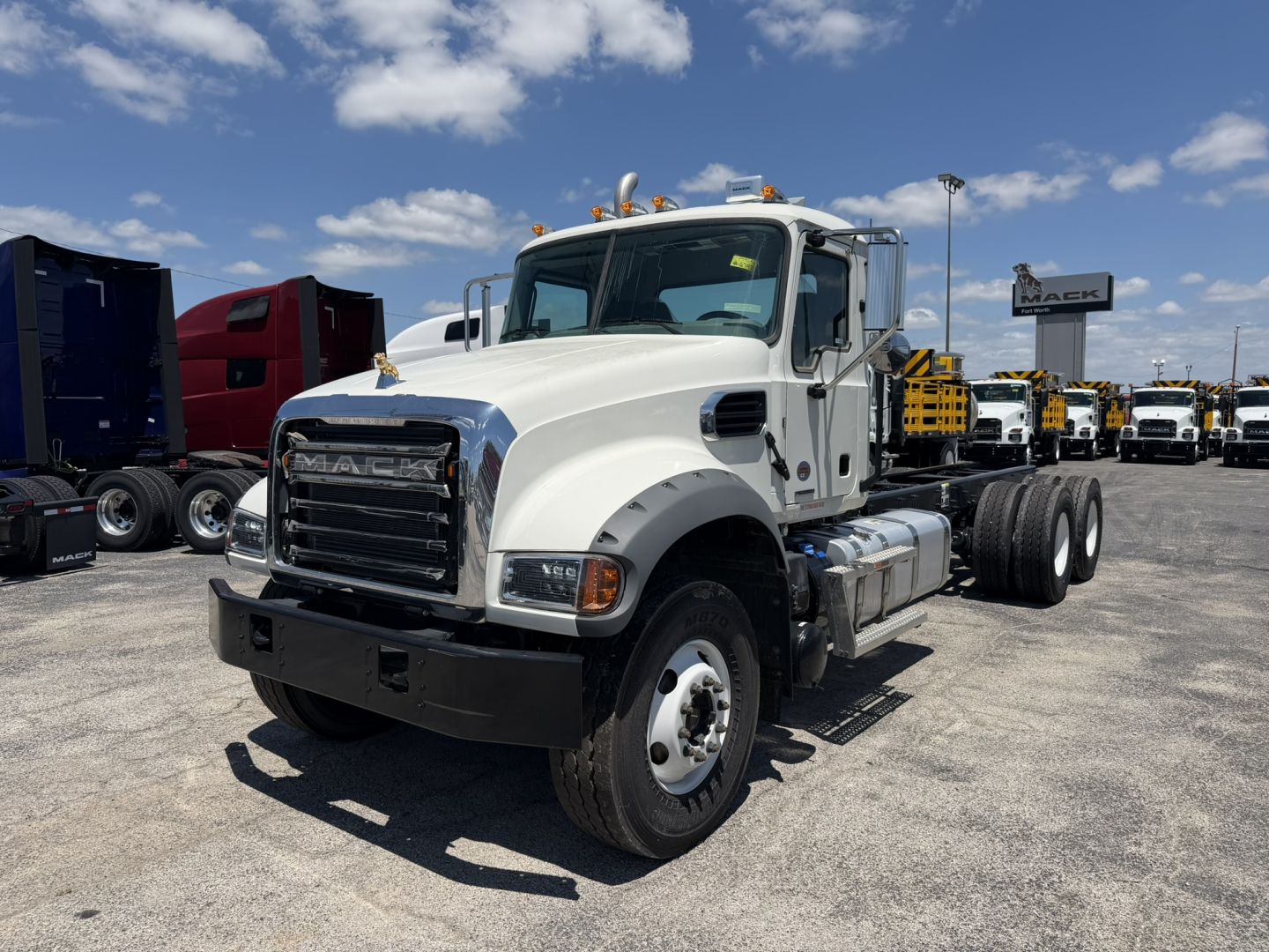 Mack GR64F cab and chassis truck in a dealership lot, white cab, dual rear axles, front grille with Mack emblem, multiple trucks in background.