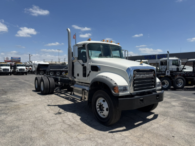 Mack GR64F truck cab and chassis in a lot, front-left view with chrome grille, dual exhaust stacks, and multiple axles in background.