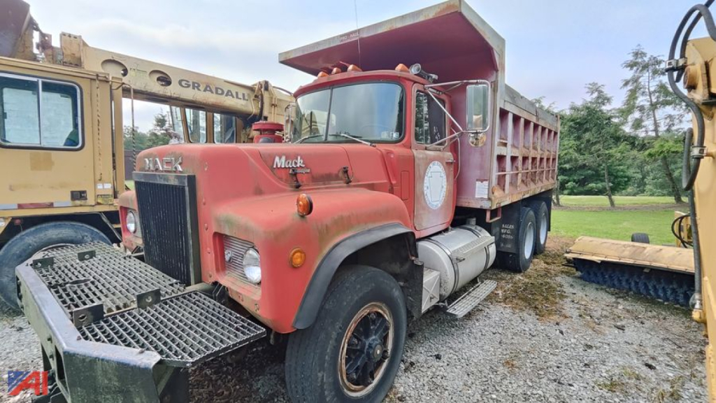 Mack RD690S dump truck in red with front grille and bumper; dump bed raised; visible text 'Mack' on hood and 'RD690S' badge; side of cab shows white circular emblem; other equipment in background.