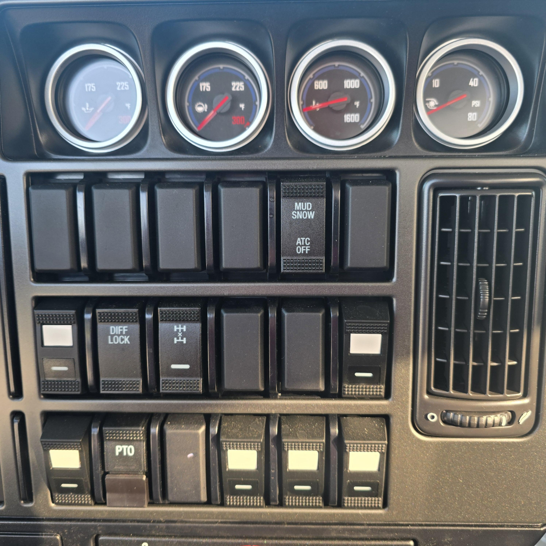 Dashboard panel of a Mack GR64B mixer showing multiple gauges and numerous switches. Gauges indicate pressure/temperature values; center stack features blank rocker switches; right side has an air vent.