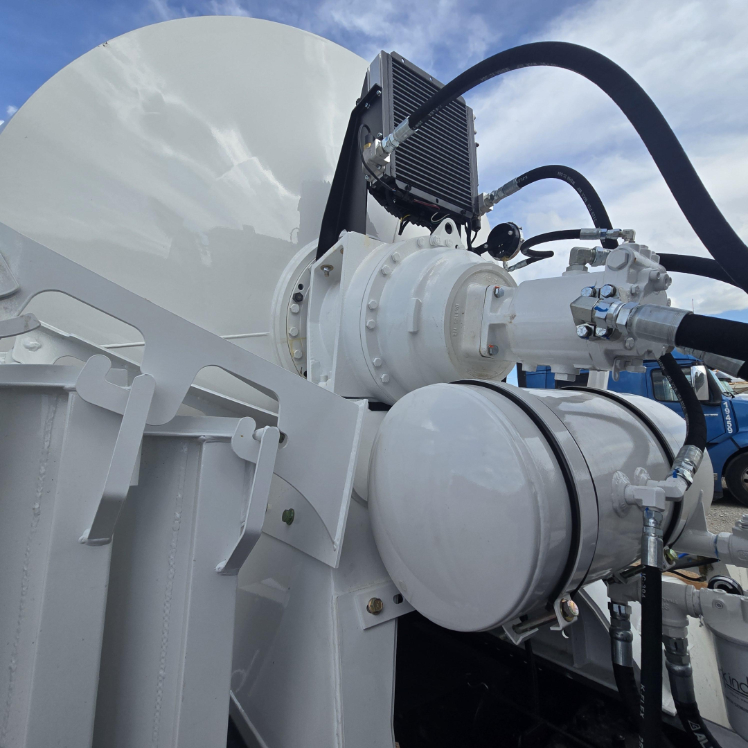 Mixer truck component close-up showing hydraulic lines, circular white valve housing, and high-pressure fittings mounted on a white tank.