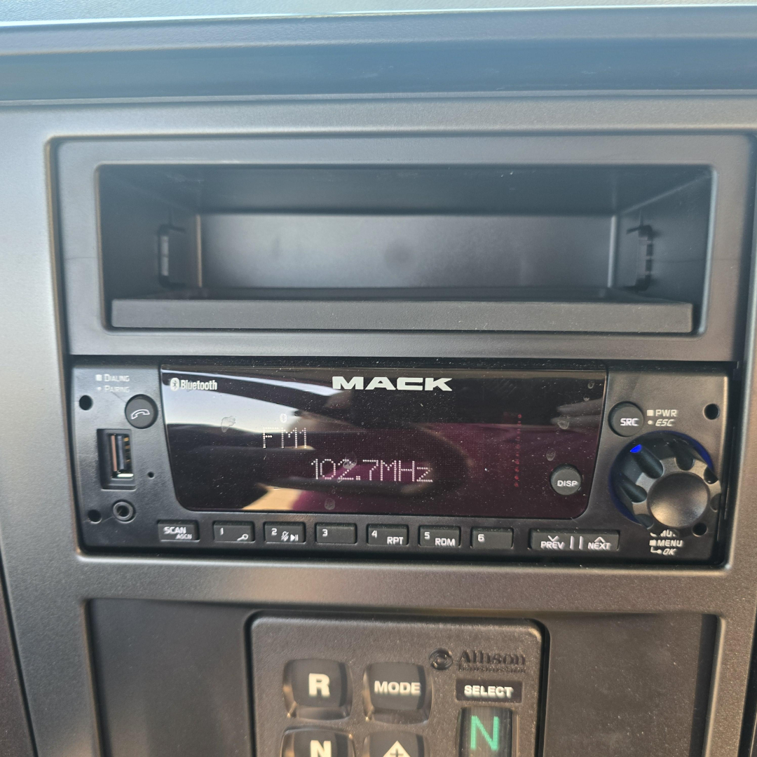 Mack radio head unit in truck cabin showing AM/FM 102.7 MHz with circular knob and USB/Bluetooth indicators.