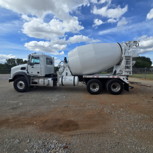 Concrete mixer truck side view showing large rotating drum, cab, and dual rear axles with white primer; environmental gravel lot and blue sky.