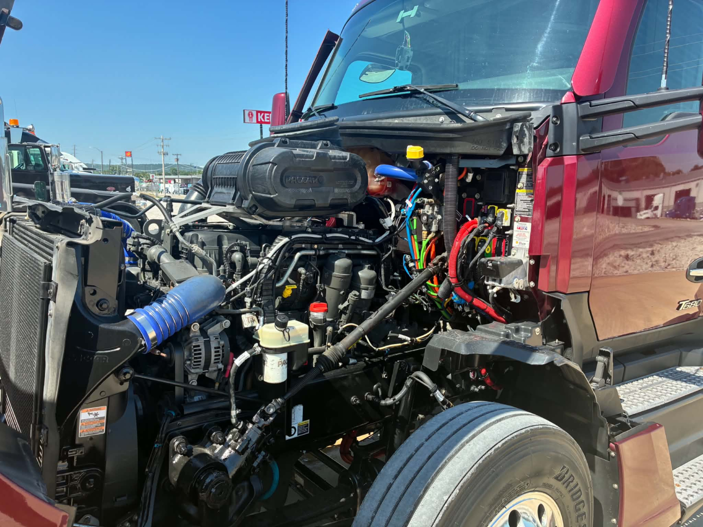 Kenworth T680 engine bay with exposed components, visible wiring harnesses, hoses, and air intake; front wheel and radiator/am radiator area visible.