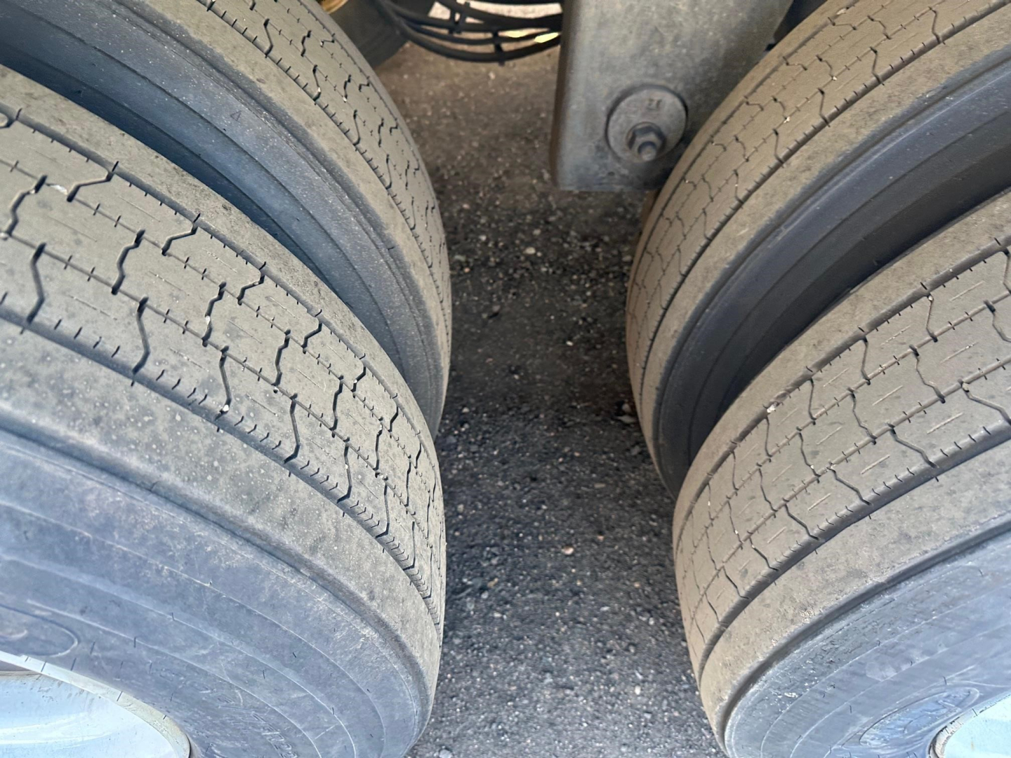 Trailer axles with dual tires viewed from between wheels; concrete surface below, part of undercarriage visible.