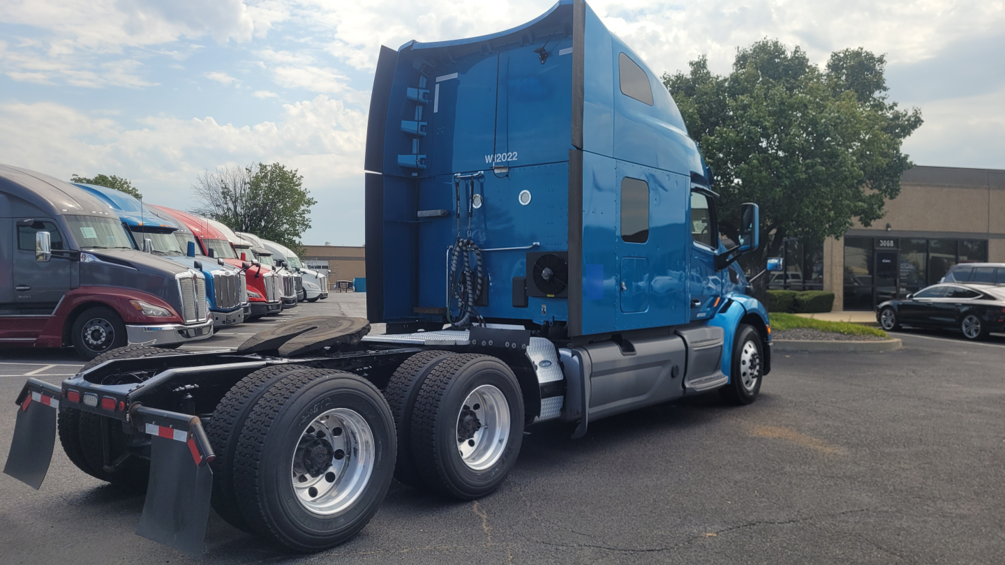 Truck rear-left side of a blue Peterbilt 579 sleeper cab tractor showing dual rear axles and fifth wheel, with other trucks in background.