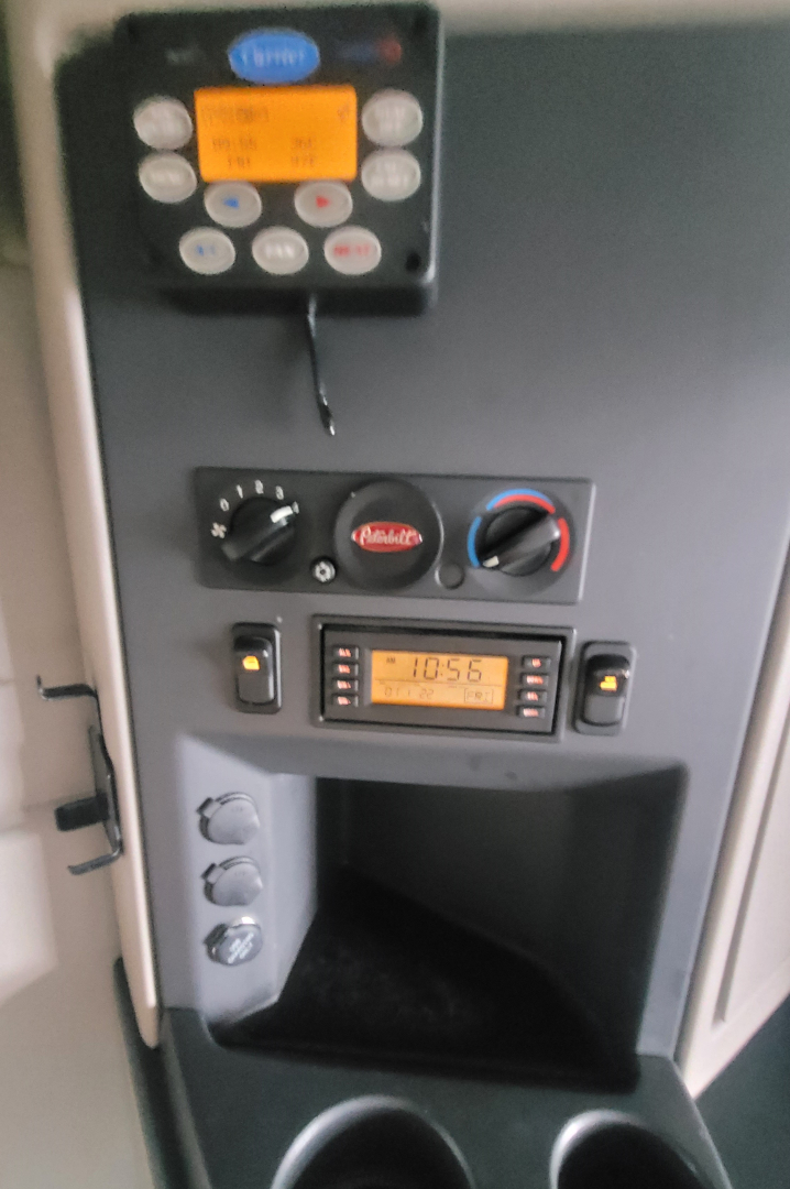 Interior cab panel of a Peterbilt 579 showing control panel with digital readouts, climate controls, and a small display; includes a small orange screen and circular knobs.