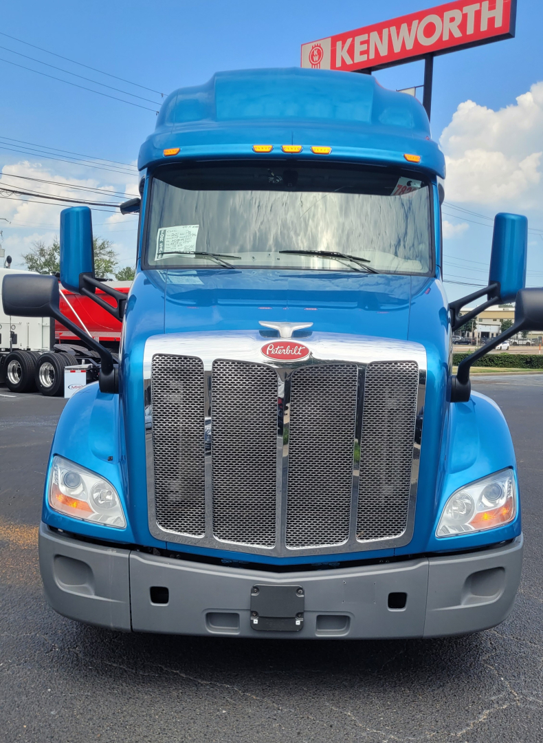 Peterbilt 579 truck front view in blue with Kenworth sign in background. Grill with Peterbilt emblem, large chrome grille, headlights, black bumper, hood and mirrors visible.