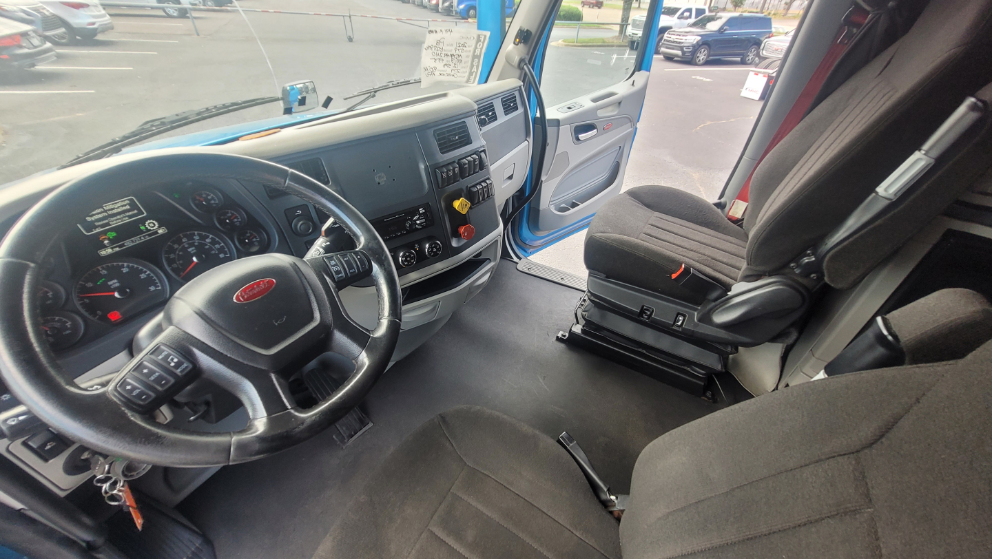Peterbilt truck cab interior showing steering wheel, dash gauges, center console, and driver seat with seatbelt; blue exterior visible through open door.