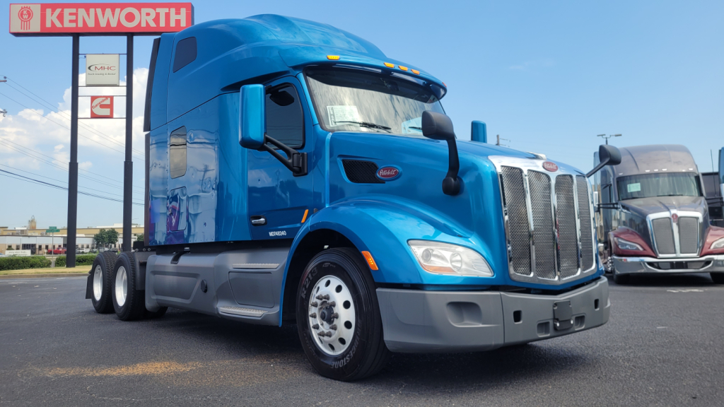 Peterbilt 579 conventional sleeper truck in blue, front-left view, parked at dealership lot with Kenworth sign in background.