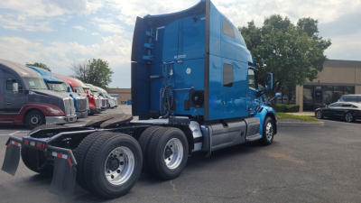 Truck rear-left side of a blue Peterbilt 579 sleeper cab tractor showing dual rear axles and fifth wheel, with other trucks in background.