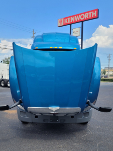 Peterbilt 579 hood front view showing blue tractor with Kenworth sign in background.