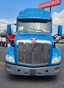 Peterbilt 579 truck front view in blue with Kenworth sign in background. Grill with Peterbilt emblem, large chrome grille, headlights, black bumper, hood and mirrors visible.