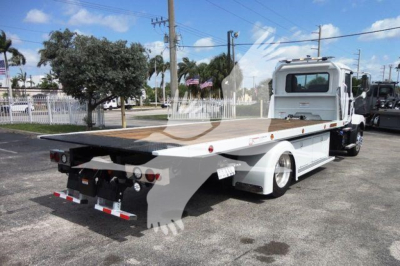 Flatbed rollback tow truck on a lot with white cab and long wooden deck; rear view shows towing equipment and lights.