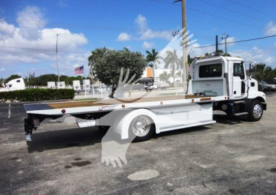 Flatbed tow truck on dealership lot with Mack MD6 cab and rollback bed, side view showing flat platform and wheel fenders.