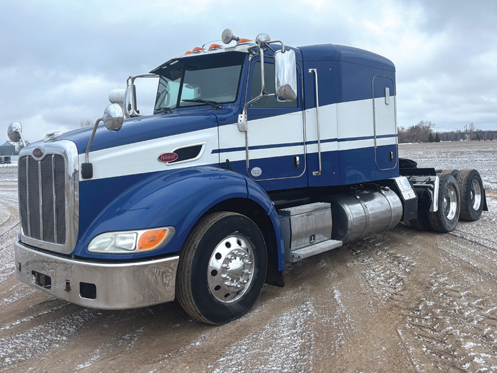 Peterbilt 384 conventional sleeper tractor in blue and white, front left view showing chrome grille, dual rear axles, fuel tanks, and side steps.