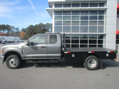 Front half of a silver 2026 Ford F350 Super Duty flatbed truck in dealership lot, side view showing flatbed with black stake body and dual rear wheels.