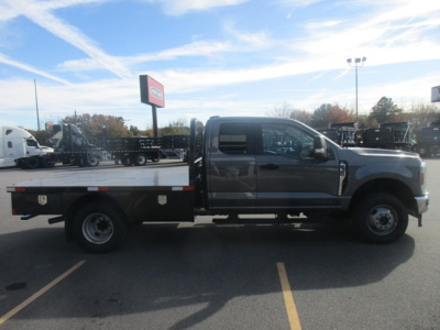 Ford F-350 Super Duty flatbed truck in a dealership lot, side view. Visible text includes Ford logo on dealership sign and a flatbed with storage compartments under the cab.