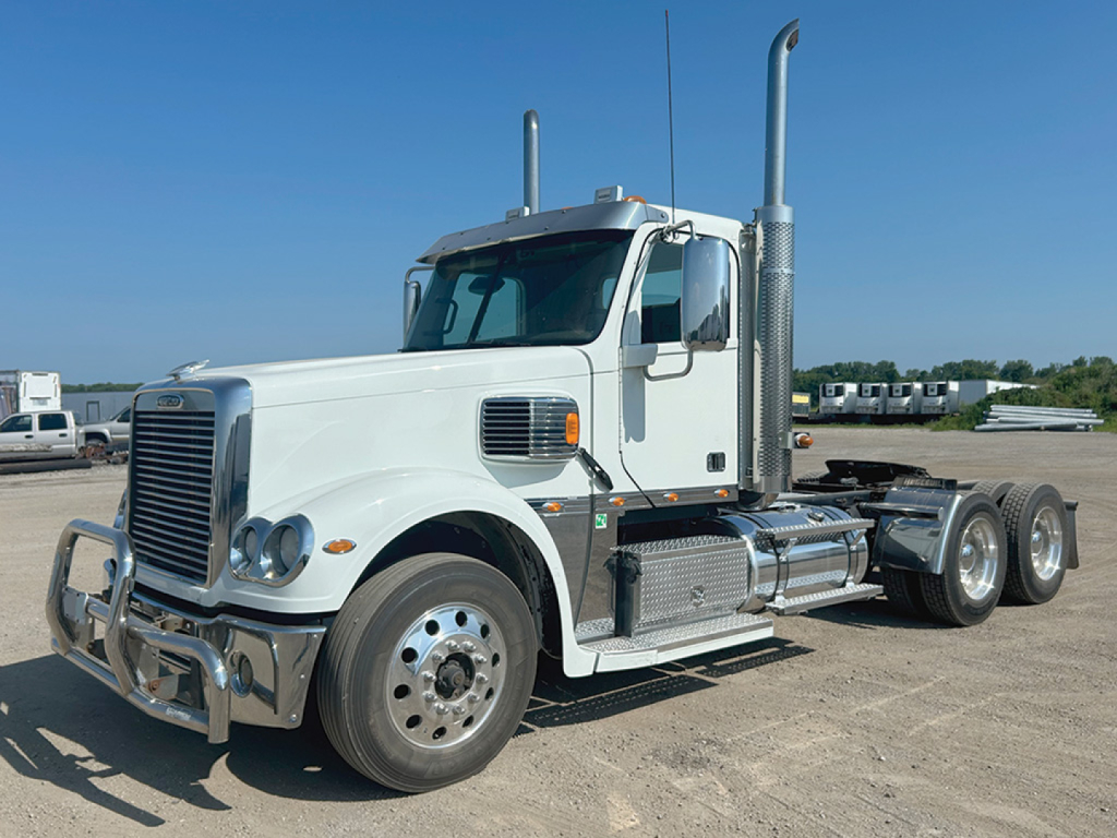 Freightliner Coronado tractor unit in light color showing front grille, dual vertical exhaust stacks, and side fuel tanks.