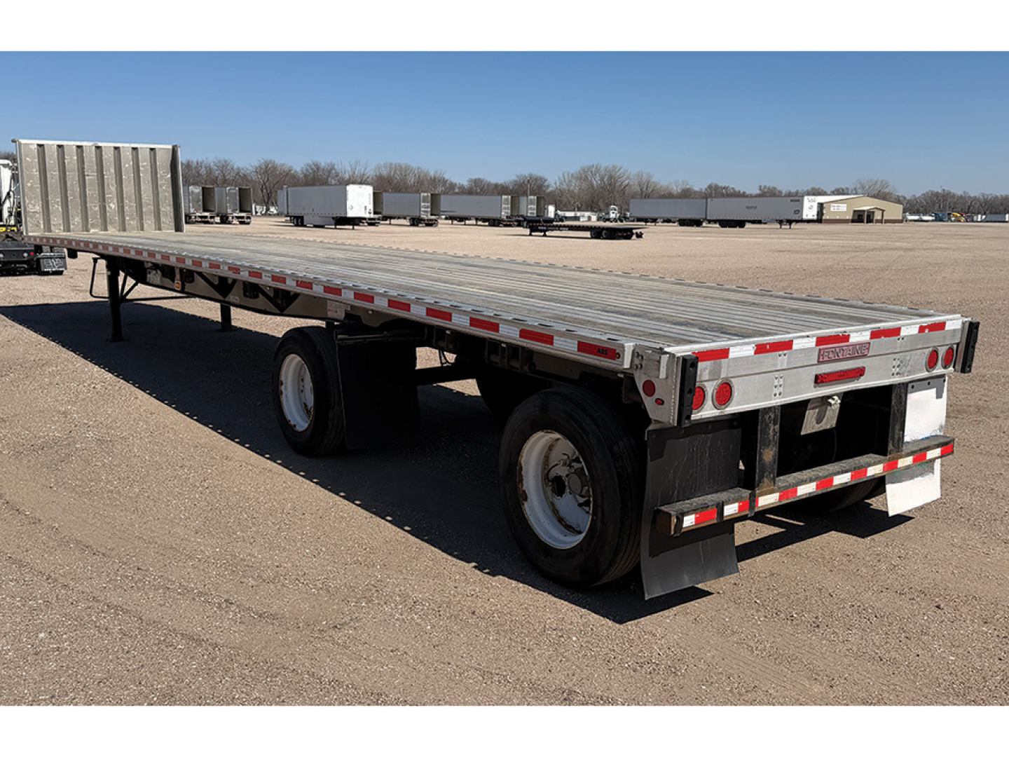 Flatbed trailer showing dual rear axles and aluminum deck with reflective tape along sides.