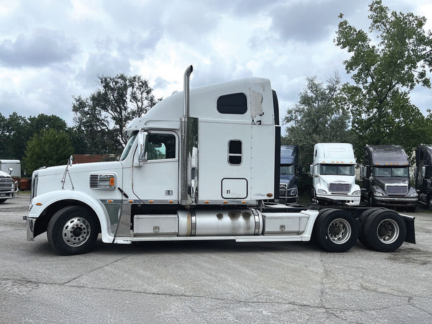 Truck cab and chassis of a white 2015 Freightliner Coronado in a lot, side view showing exhaust stacks and sleeper cab, several other trucks in background.