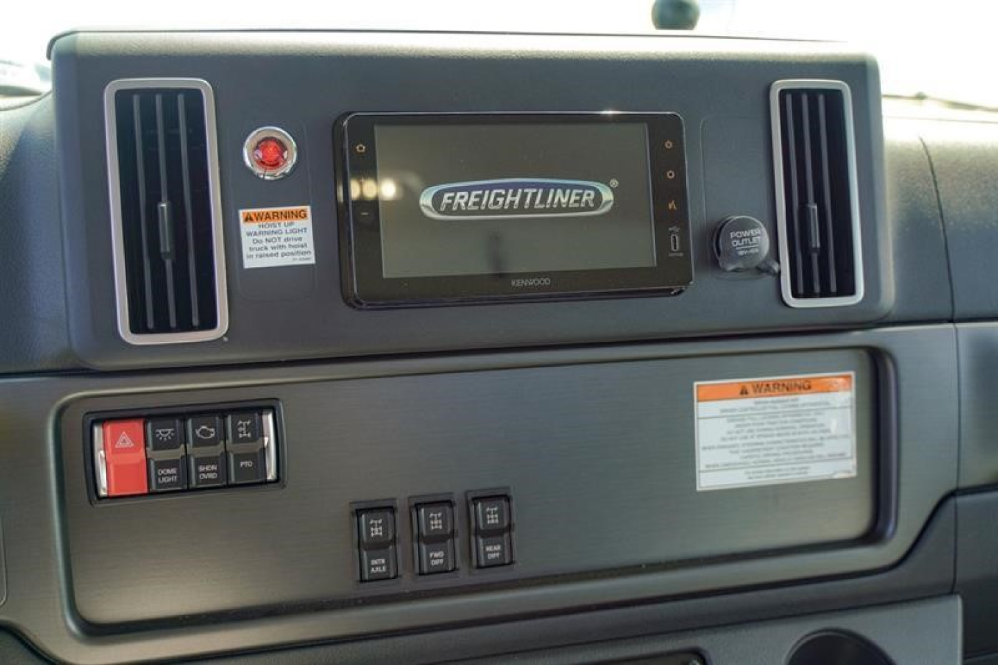 Freightliner 114SD dashboard showing Kenworth-style screen with Freightliner logo, two vertical air vents, control panel, warning labels, and knob.