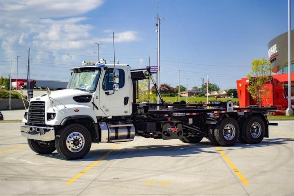Freightliner 114SD roll-off truck in showroom lot, side view, white cab, chassis with empty bed, multiple axles, blue sky.