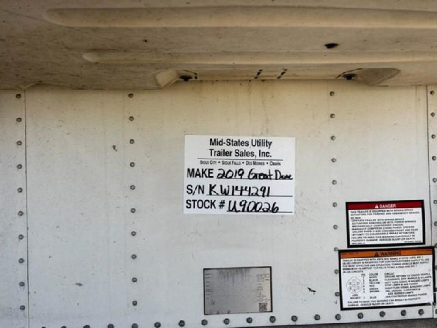 Trailer side showing stock label with Make 2019 Great Dane, S/N NKJ414? Stock # U90026, mid-states utility trailer sales placard, various caution labels.